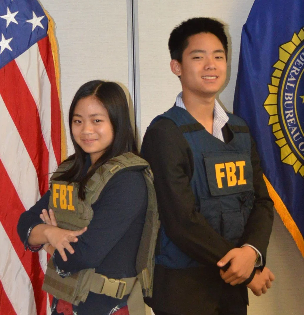 Two teenagers standing in front of U.S. flags, wearing FBI vests, smiling.