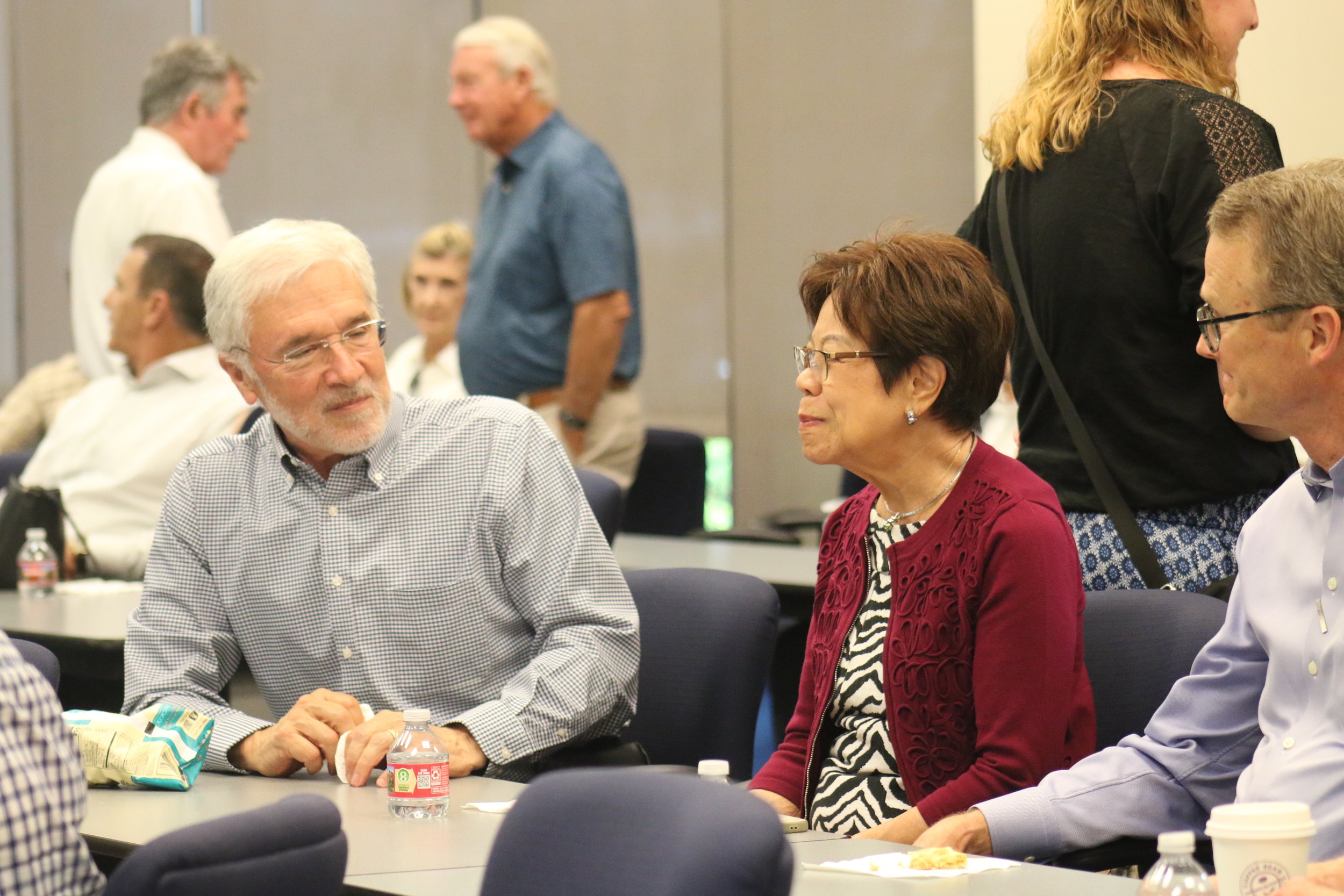 People in a conference room engaging in conversations, with some sitting at tables and others standing, eating, or talking.
