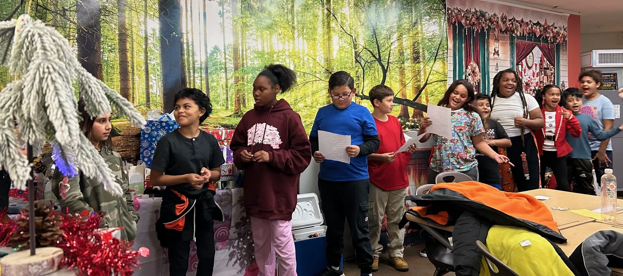 Children gathered at a holiday party, some holding papers, smiling and laughing, with a festive backdrop of woodland scene and holiday decorations.