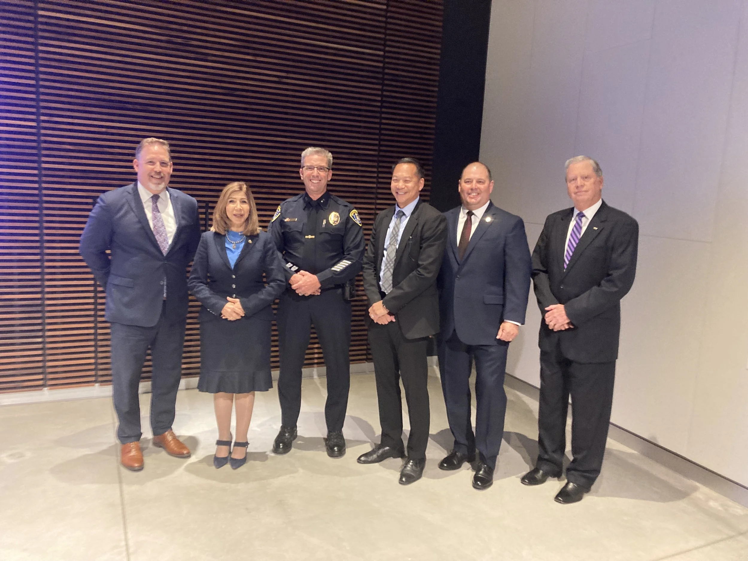 Group of seven professionally dressed people, including a police officer in uniform, standing in front of a wooden wall and grey wall on the right, smiling for a group photo.