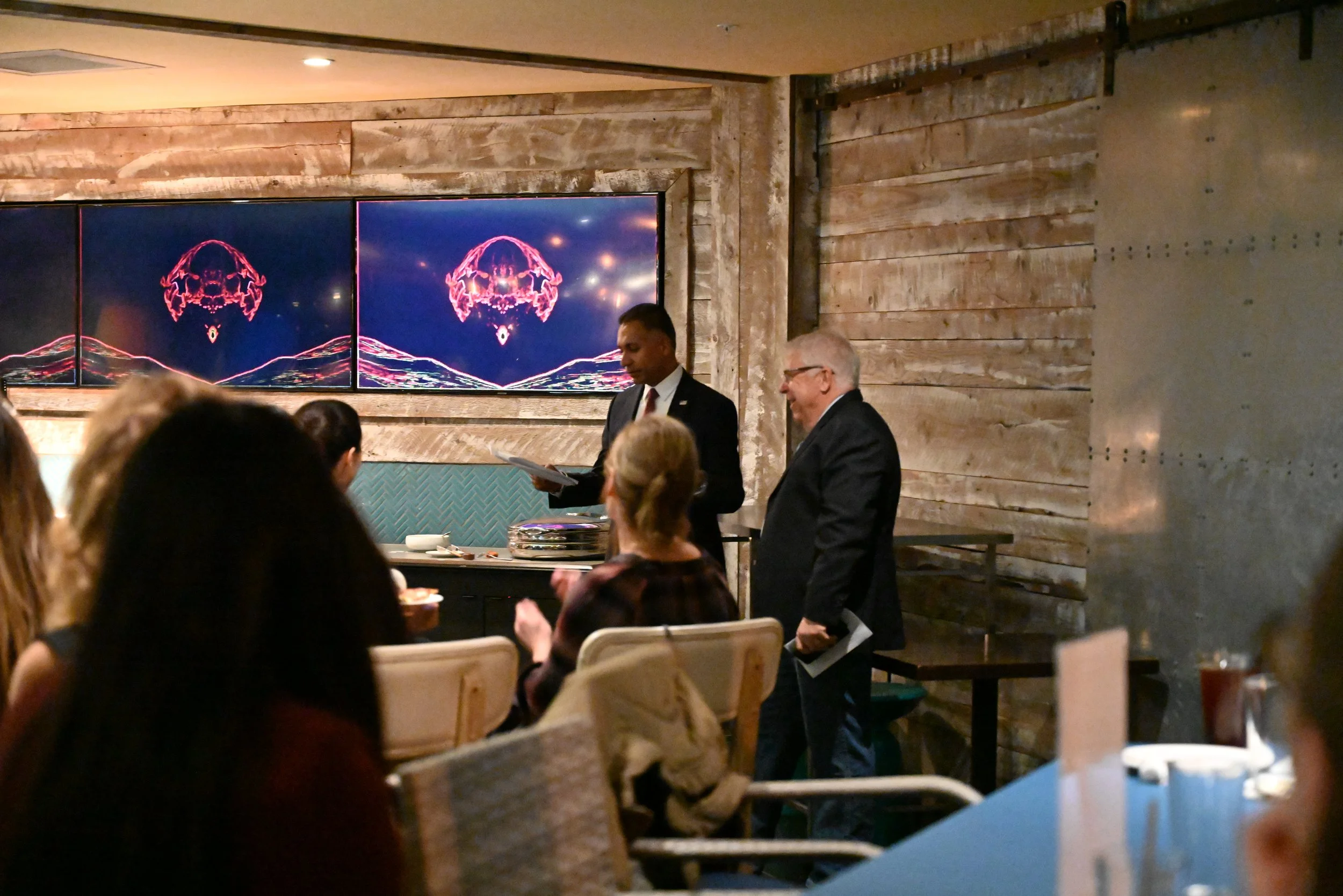 Two men in suits standing in front of a group of seated people in a rustic indoor setting. One man is holding a clipboard and the other a paper, and they appear to be speaking or presenting. Behind them, a large wall decorated with wood paneling and 
