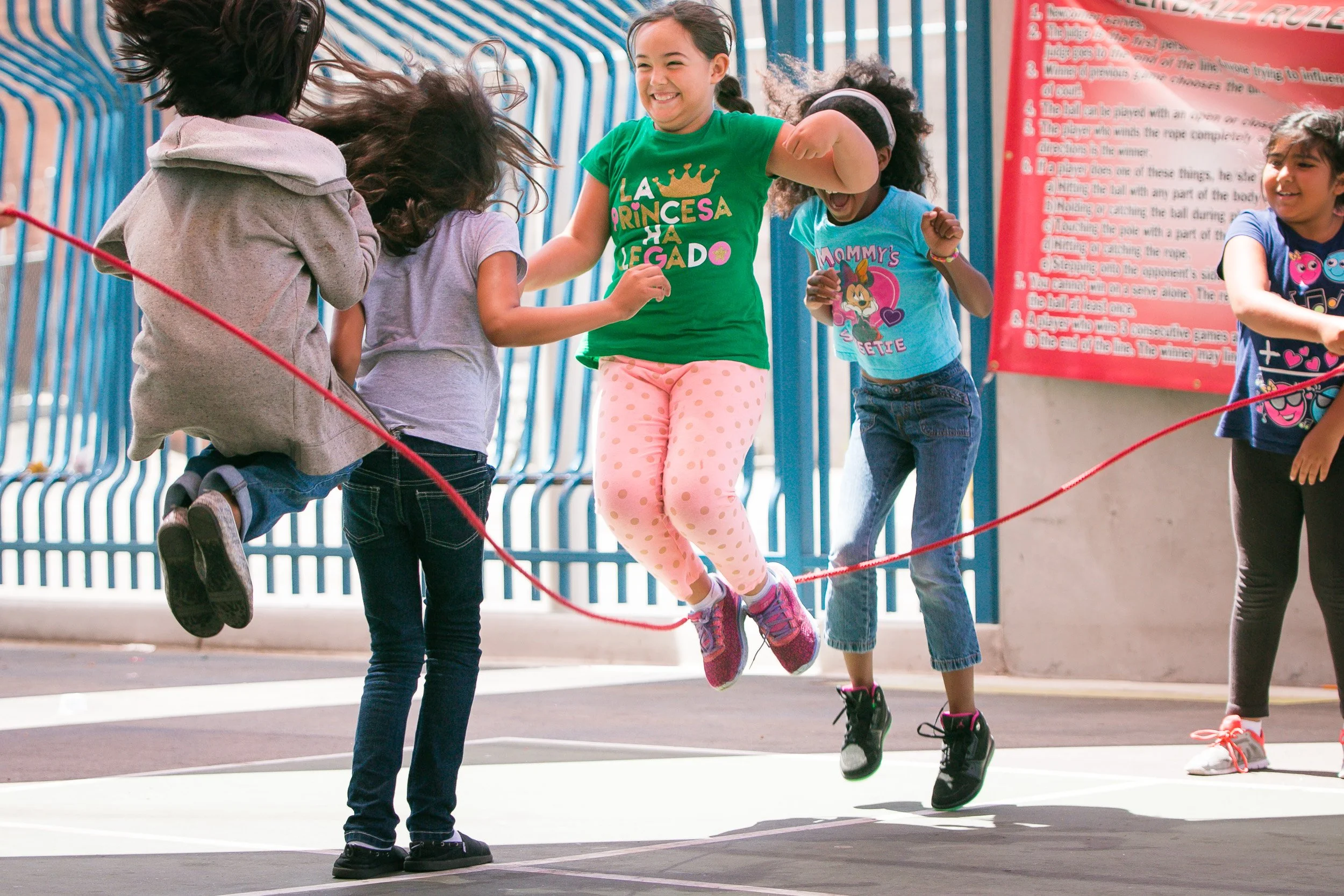 Five girls playing jump rope outdoors, one girl in mid-air, smiling, wearing a green shirt and pink polka dot leggings, others around her watching and smiling.