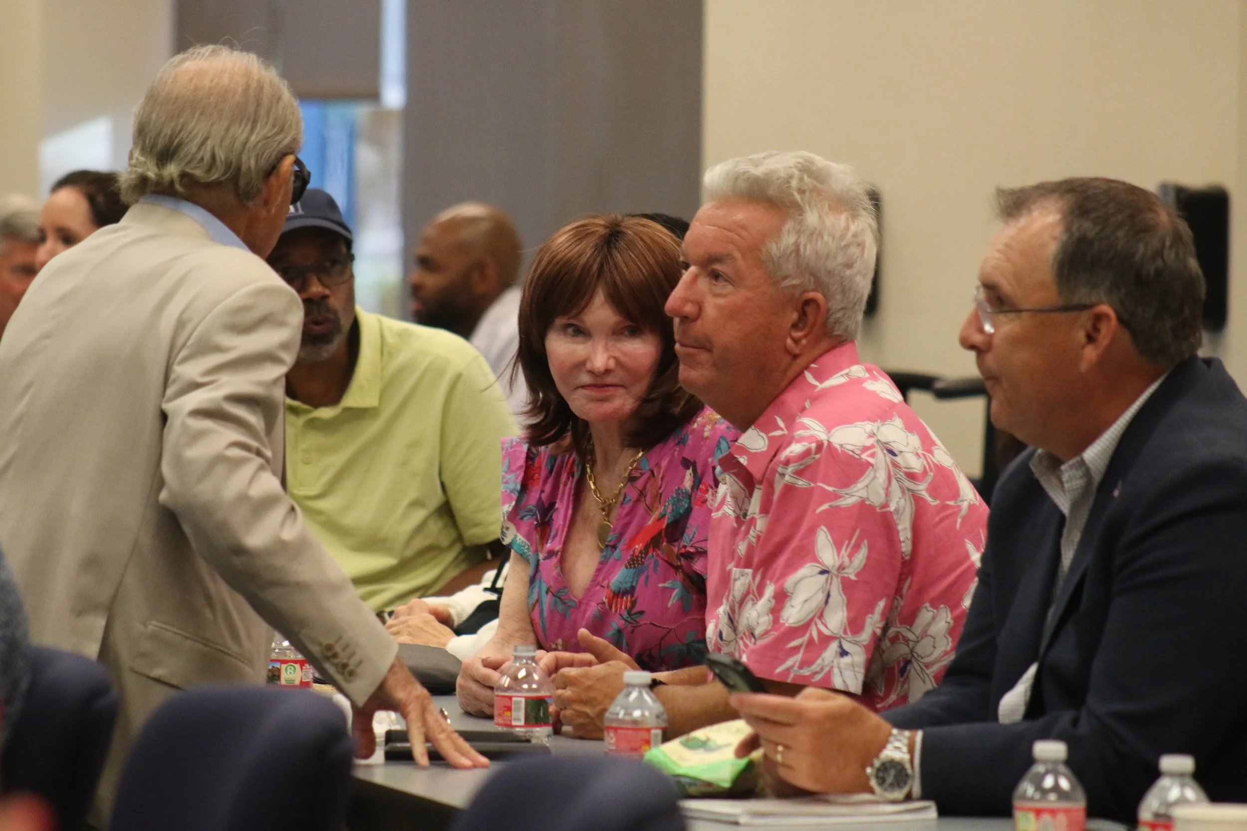 A group of adults sitting at a table in a conference room, engaged in conversation, with water bottles and papers on the table.