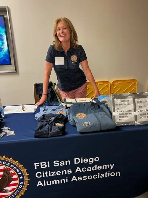 A smiling woman in a navy polo shirt stands behind a table displaying FBI San Diego Citizens Academy Alumni Association merchandise, including clothing and informational materials.