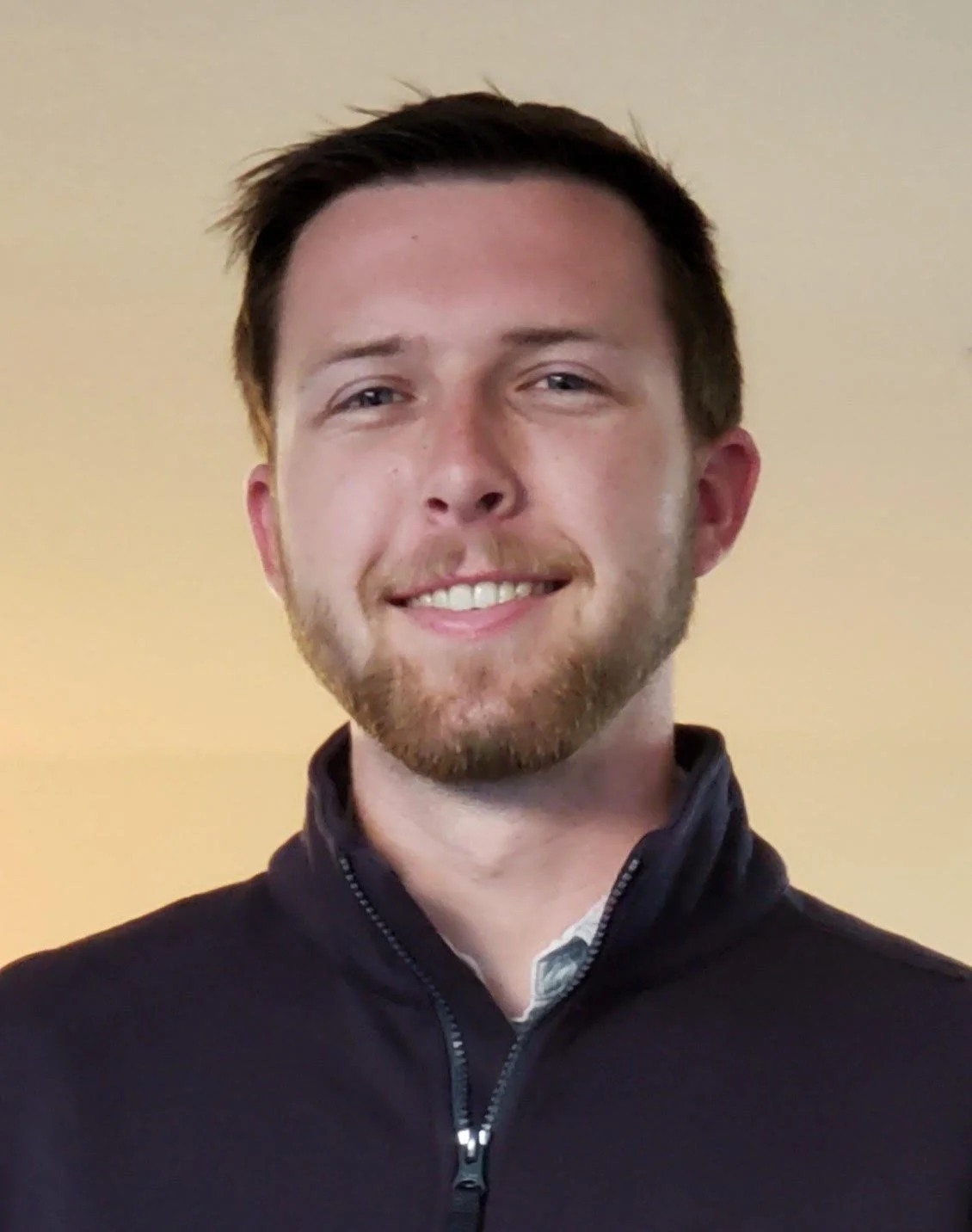 A young man with short brown hair and a beard, smiling, wearing a black zip-up jacket, standing against a yellow wall.