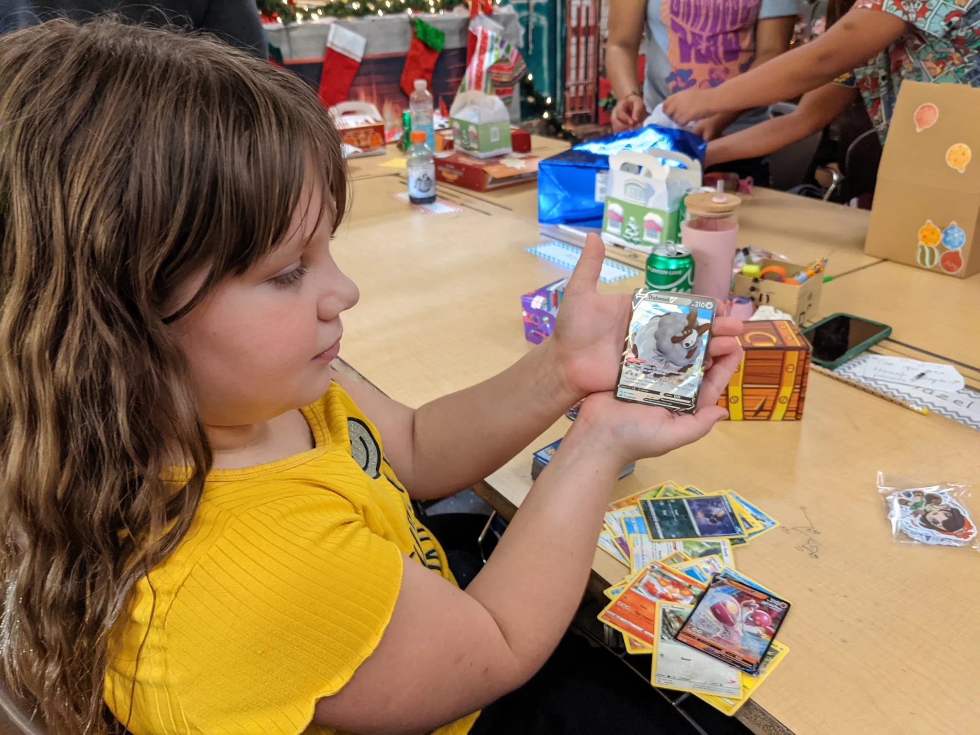 A young girl with long brown hair wearing a yellow shirt playing a trading card game at a holiday party. She is holding a card with a bear on it and looking at her cards on the table. The table is filled with Pokémon cards and Christmas-themed gift b