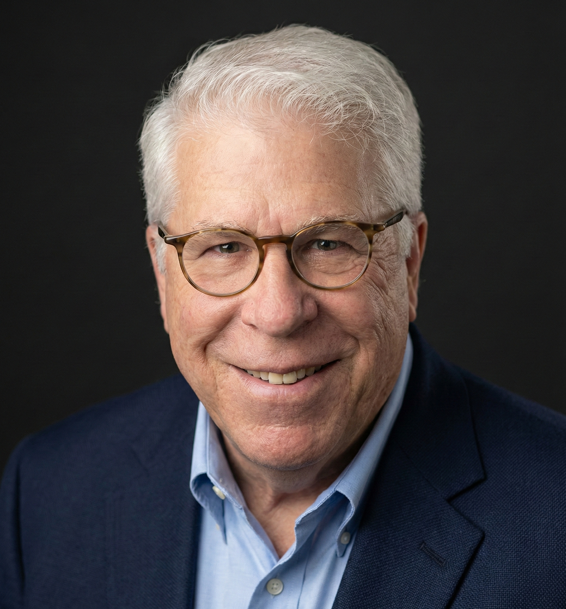 A close-up portrait of an older man with gray hair, glasses, and a serious expression, wearing a dark suit and light blue shirt against a dark background.