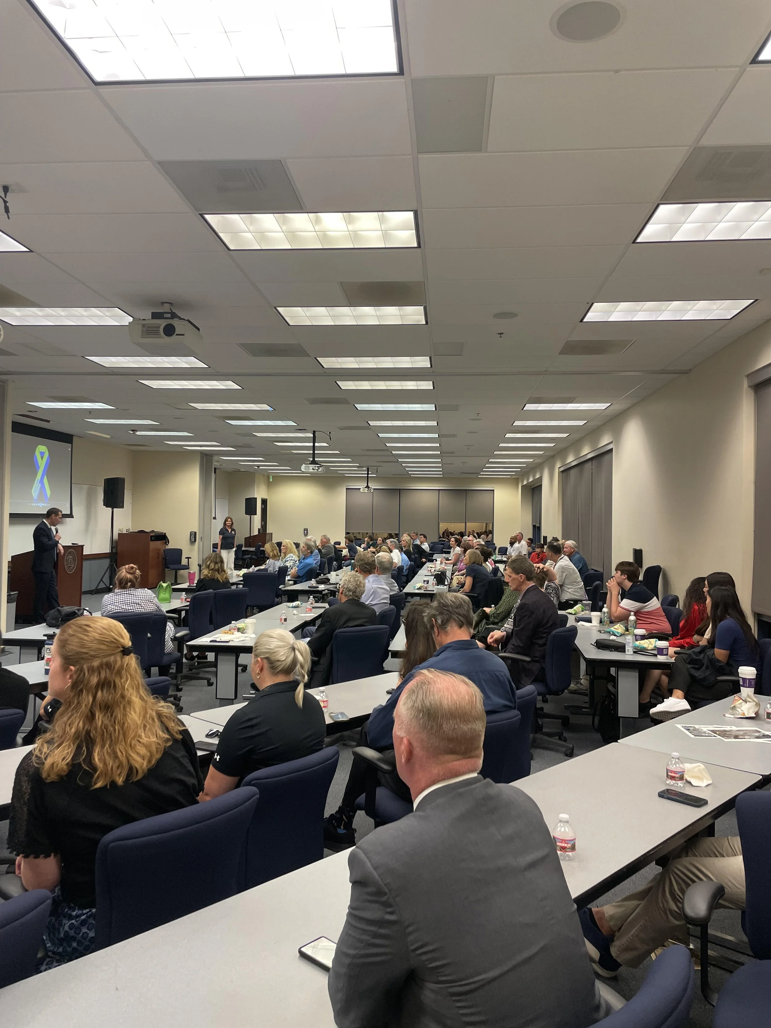 A large conference room filled with attendees listening to a speaker at the front. People are seated at long tables with notebooks and bottled water. A slide with a ribbon graphic is displayed on a projector screen.