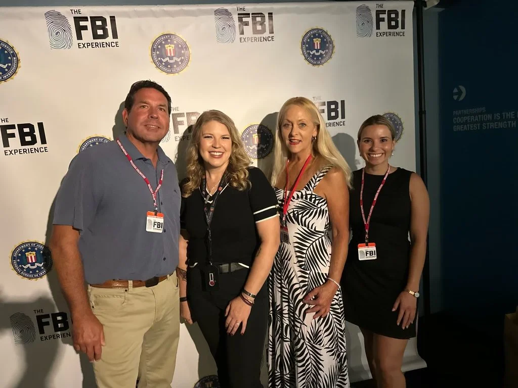 Four people posing in front of an FBI backdrop at an event, three women and one man, all wearing event badges.