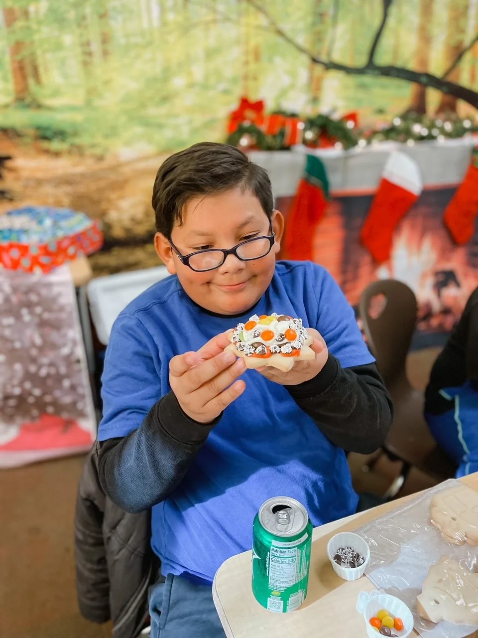 A boy with glasses wearing a blue shirt and black long-sleeve shirt underneath, holding a decorated cookie with orange, white, and black icing, at a Christmas-themed party with stockings and a fireplace decoration in the background.