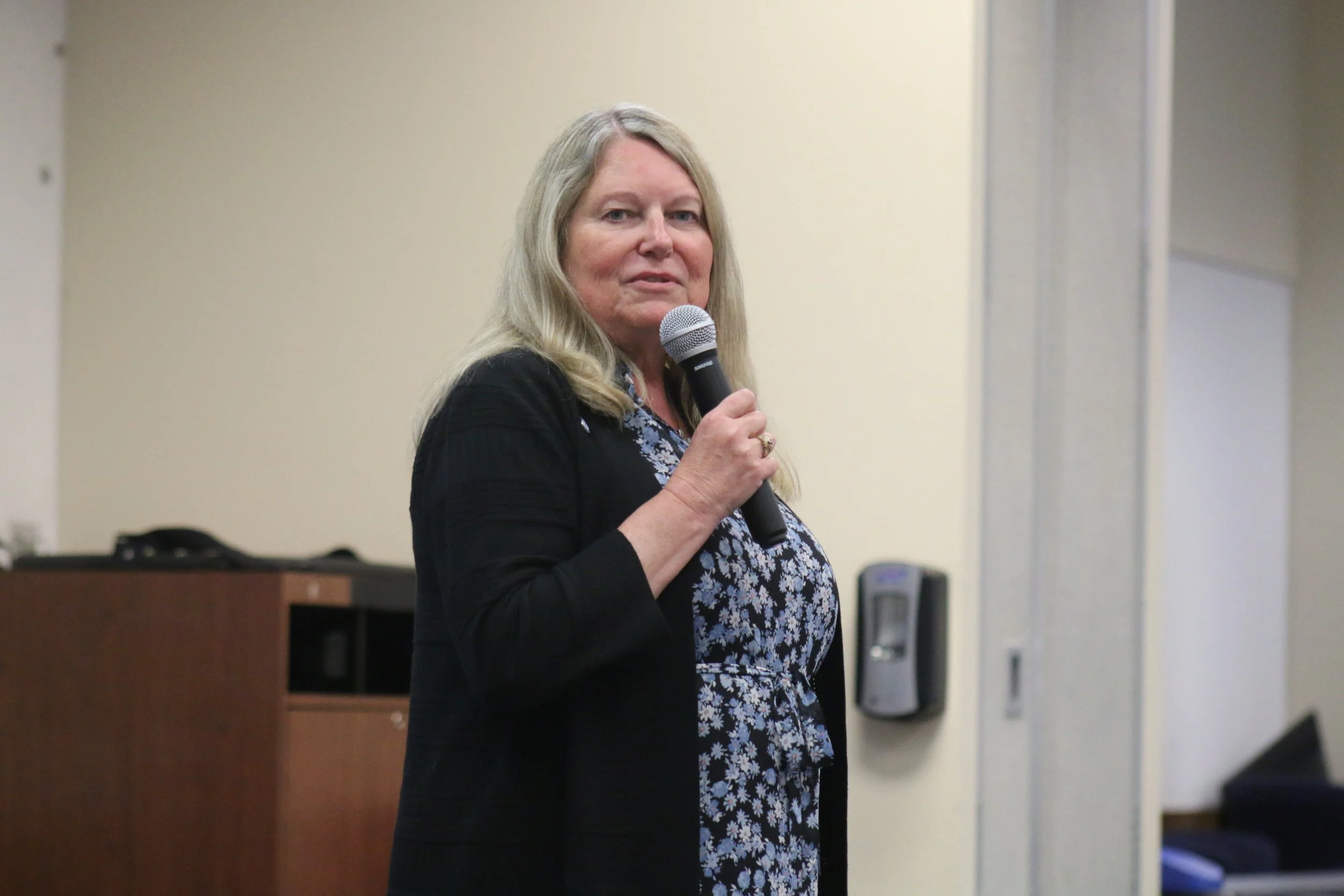 A woman with long blonde hair holding a microphone, wearing a blue floral dress and a black cardigan, standing indoors.