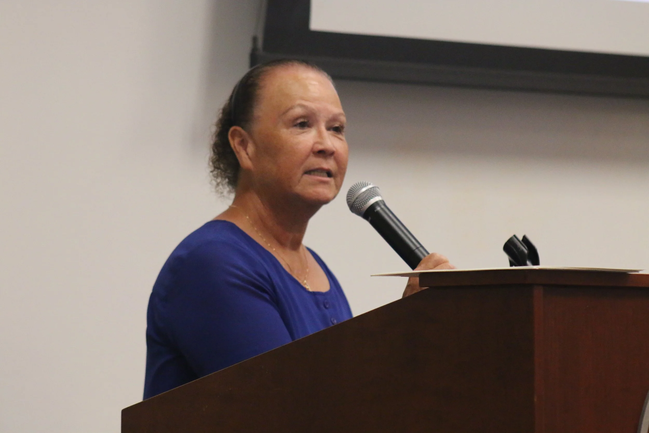 A woman with short curly hair, wearing a royal blue blouse, speaking into a microphone at a podium in a conference room.