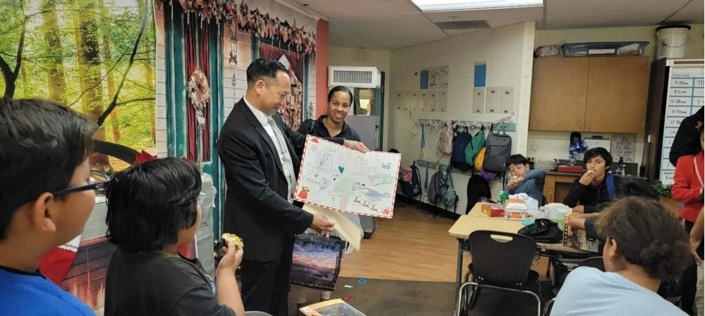 A group of school students in a classroom, with one man in a suit and a woman holding a large decorated poster board, engaging with the students.
