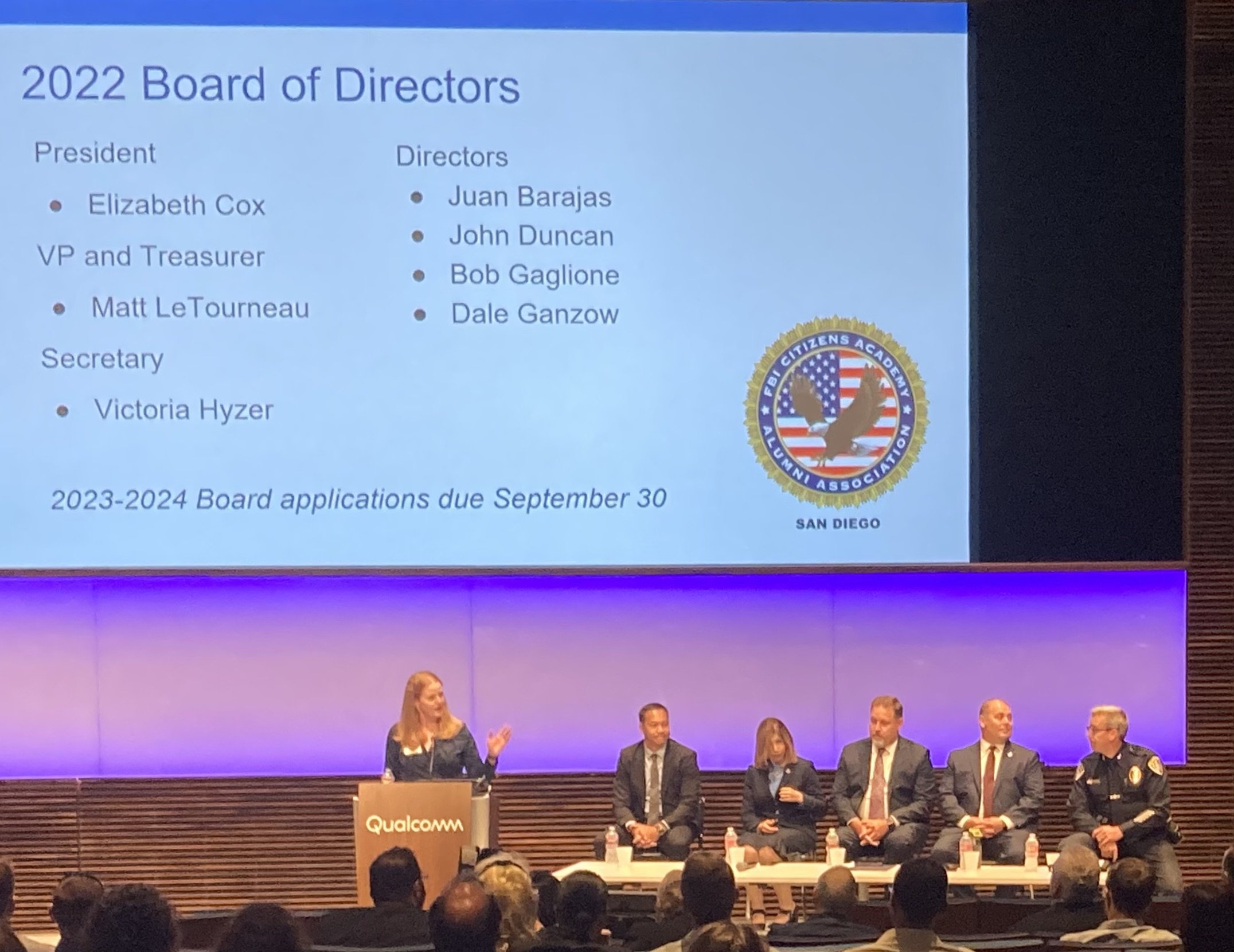 Panel of five people seated at a table with a woman standing at a podium labeled Qualcomm, discussing 2022 Board of Directors for the KEI Citizens Academy Alumni Association, with a large screen showing the board members' names and titles.