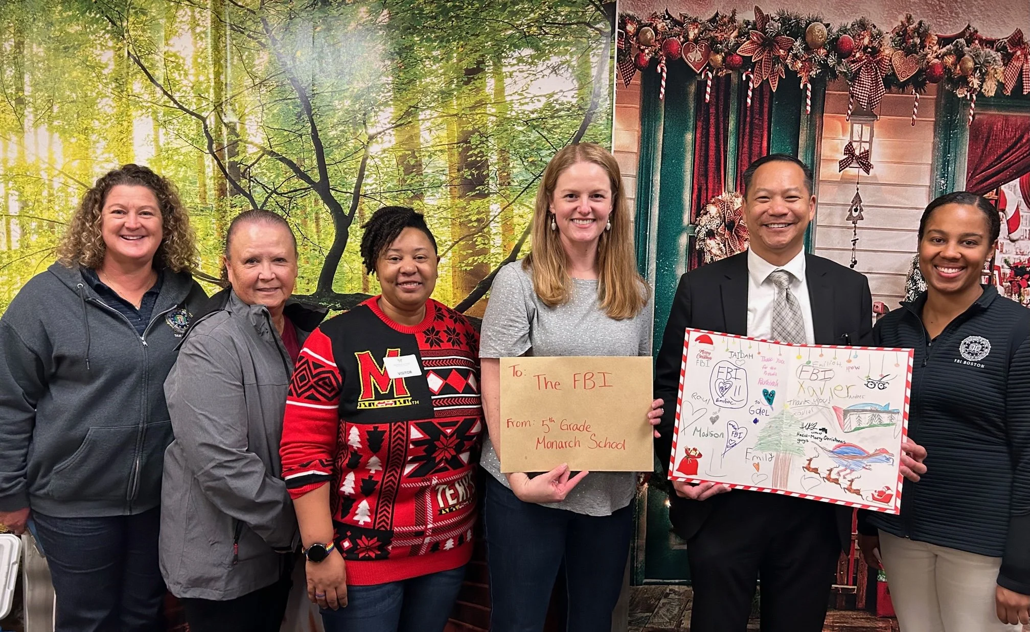 Group of seven people standing indoors, holding signs and artwork, with a holiday and nature-themed background.