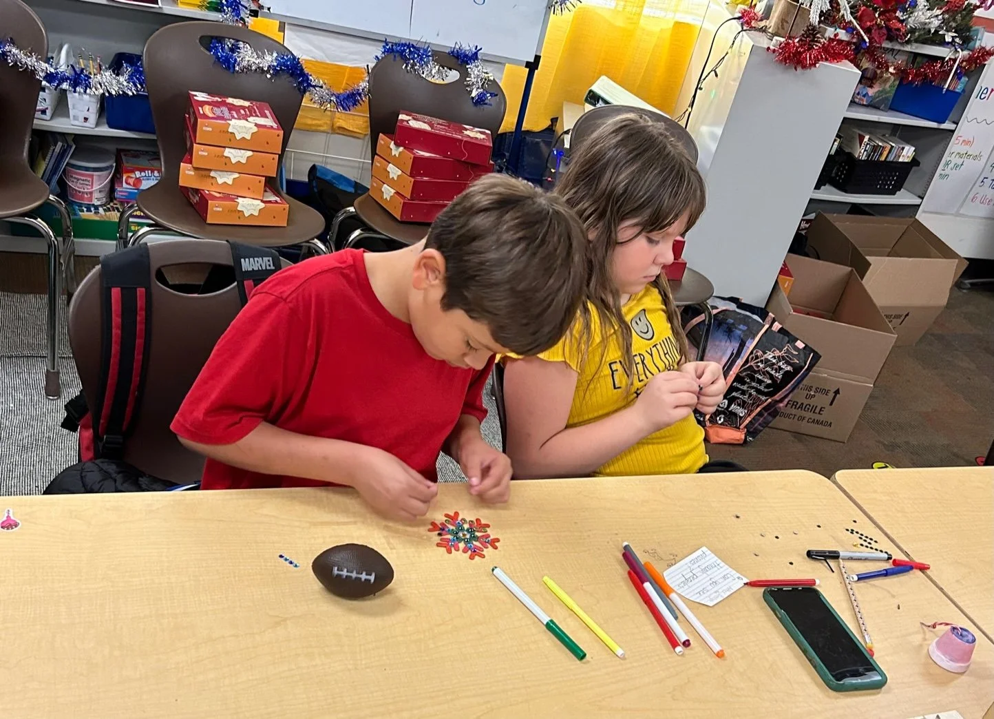 Two children sitting at a table in a classroom, decorating Christmas ornaments with markers and stickers, with holiday decorations and boxes in the background.