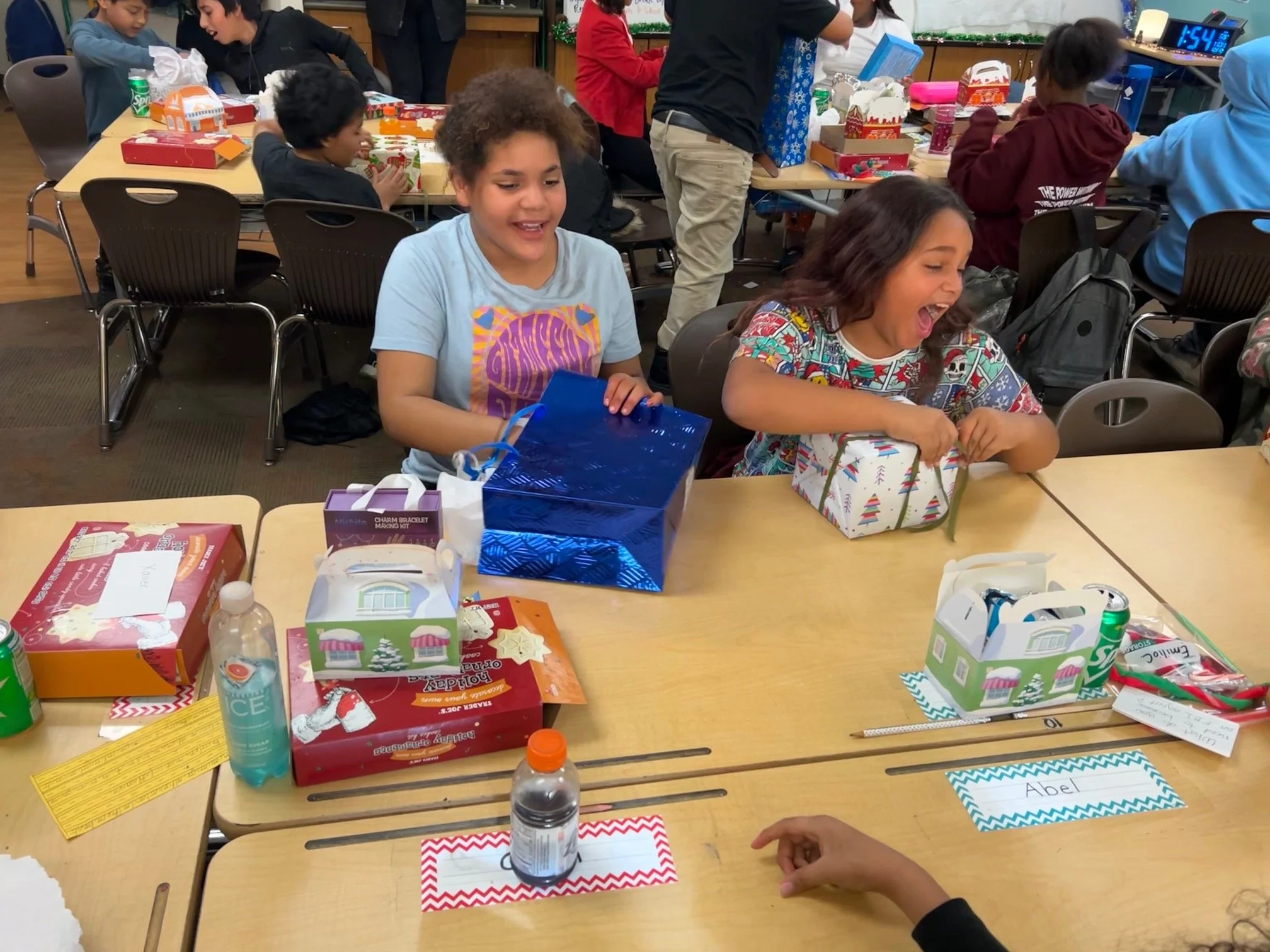 Two girls sitting at a table opening gifts with joyful expressions, surrounded by other children and holiday-themed presents.