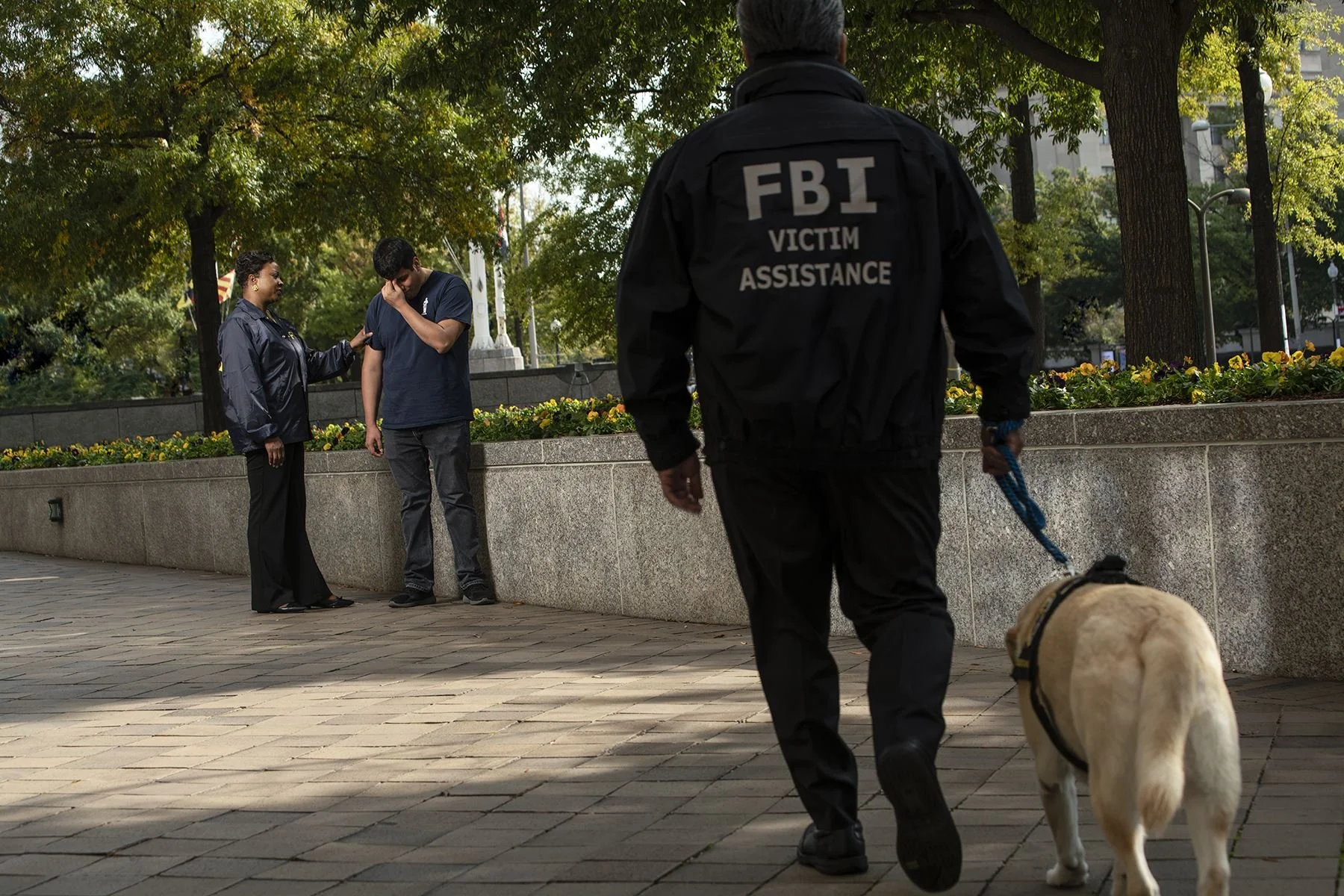 FBI Victim Assistance agent walking a service dog in front of a memorial park, where a woman and a man are in a moment of mourning or reflection.