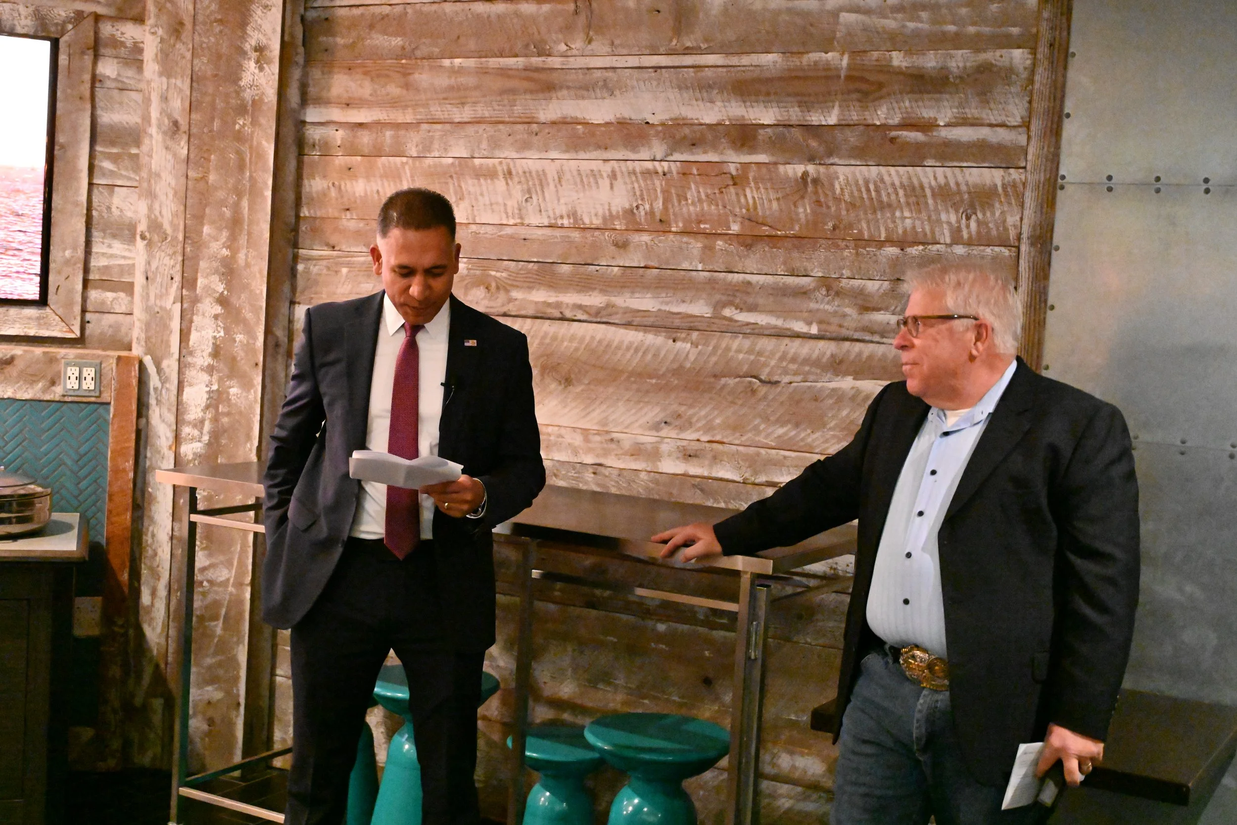 Two men in suits having a conversation indoors, one is holding papers and the other has a microphone, with a rustic wooden wall background.