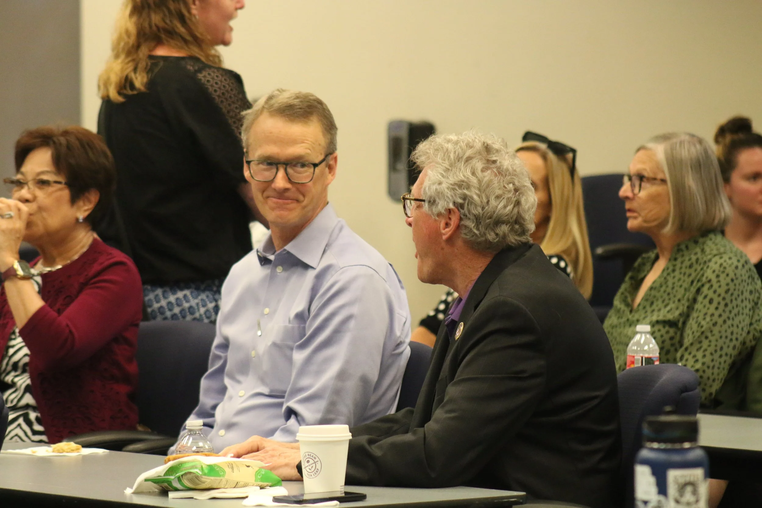 People sitting in a conference room, engaging in discussion. A man in a blue shirt and glasses looks toward the camera with a slight smile. Other individuals, including women wearing glasses and patterned clothing, are seated or standing nearby, some