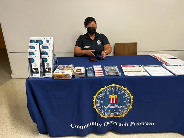 A woman wearing a black face mask and dark shirt sits behind a table covered with a blue cloth bearing the Department of Justice emblem and 'Community Outreach Program' text, with informational pamphlets and promotional items displayed.