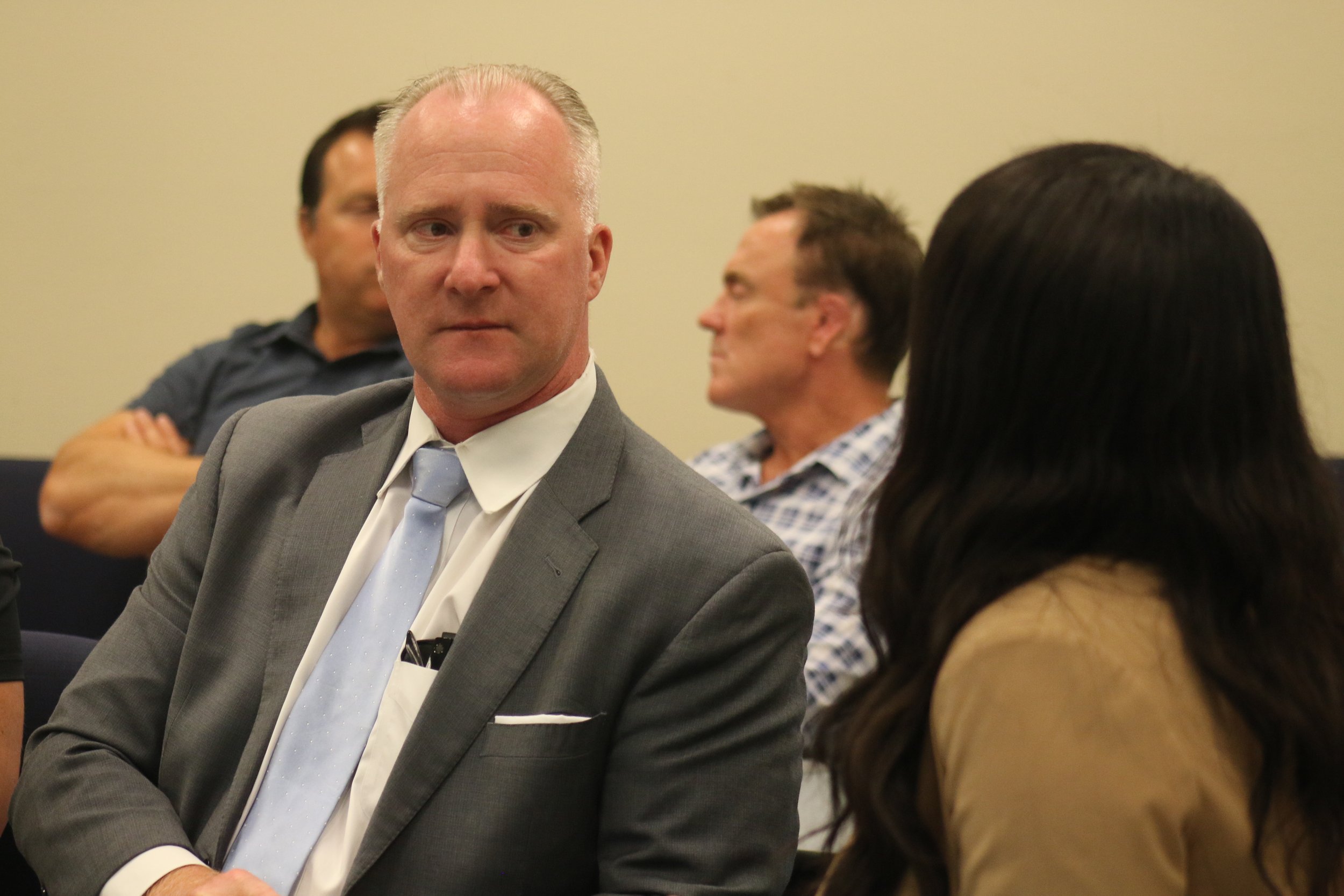 A group of people sitting in a room, with a man in a gray suit and light blue tie in focus, looking slightly to his left. Other people are blurred in the background.