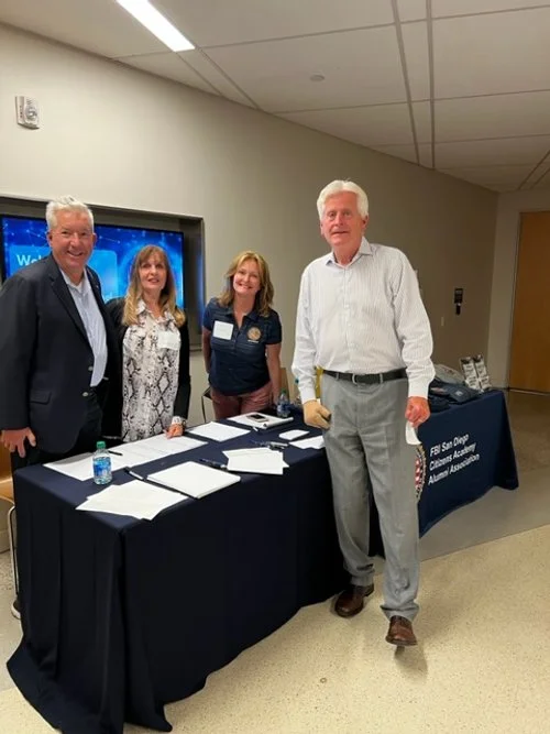 Four people standing behind a table with papers, water bottles, and a sign that reads 'FBI San Diego California Alumni Association' in an indoor setting.