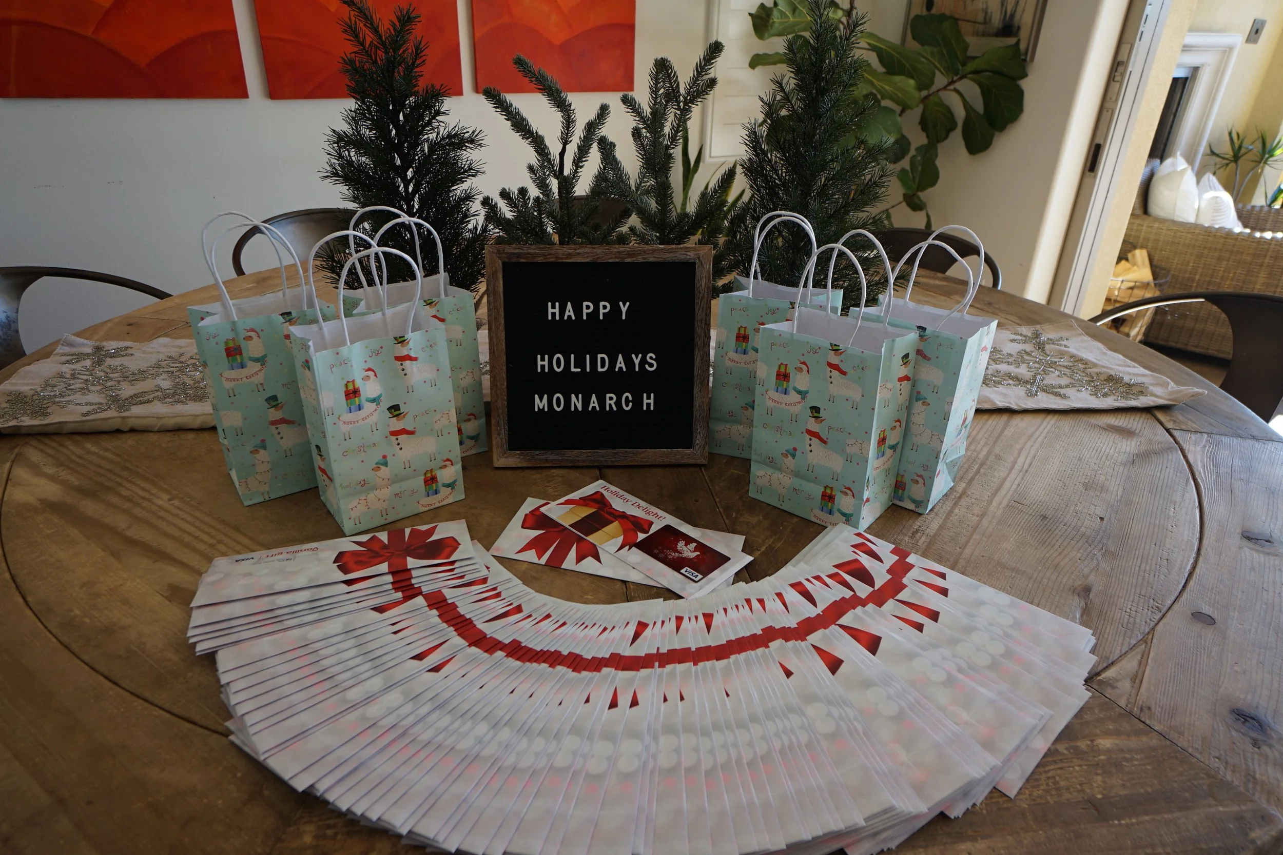 A dining table decorated for the holidays with gift bags, holiday cards, and a small blackboard sign that reads 'HAPPY HOLIDAYS MONARCH'. There are three small Christmas trees in the background and holiday-themed decorations on the table.