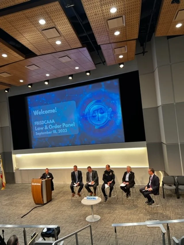 Panel of six people seated on stage in front of a large screen displaying a welcome message for a law and order panel discussion, September 18, 2023.
