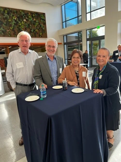 Four older adults, two men and two women, standing around a high-top table at a social event indoors, with plates, beverages, and name tags, in a modern building with large windows and greenery decor on the wall.