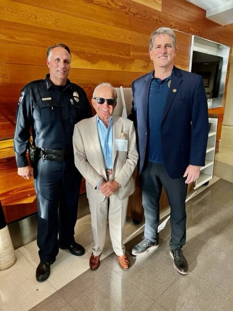 Three men standing together indoors, two in police uniforms and one in a beige suit and sunglasses.