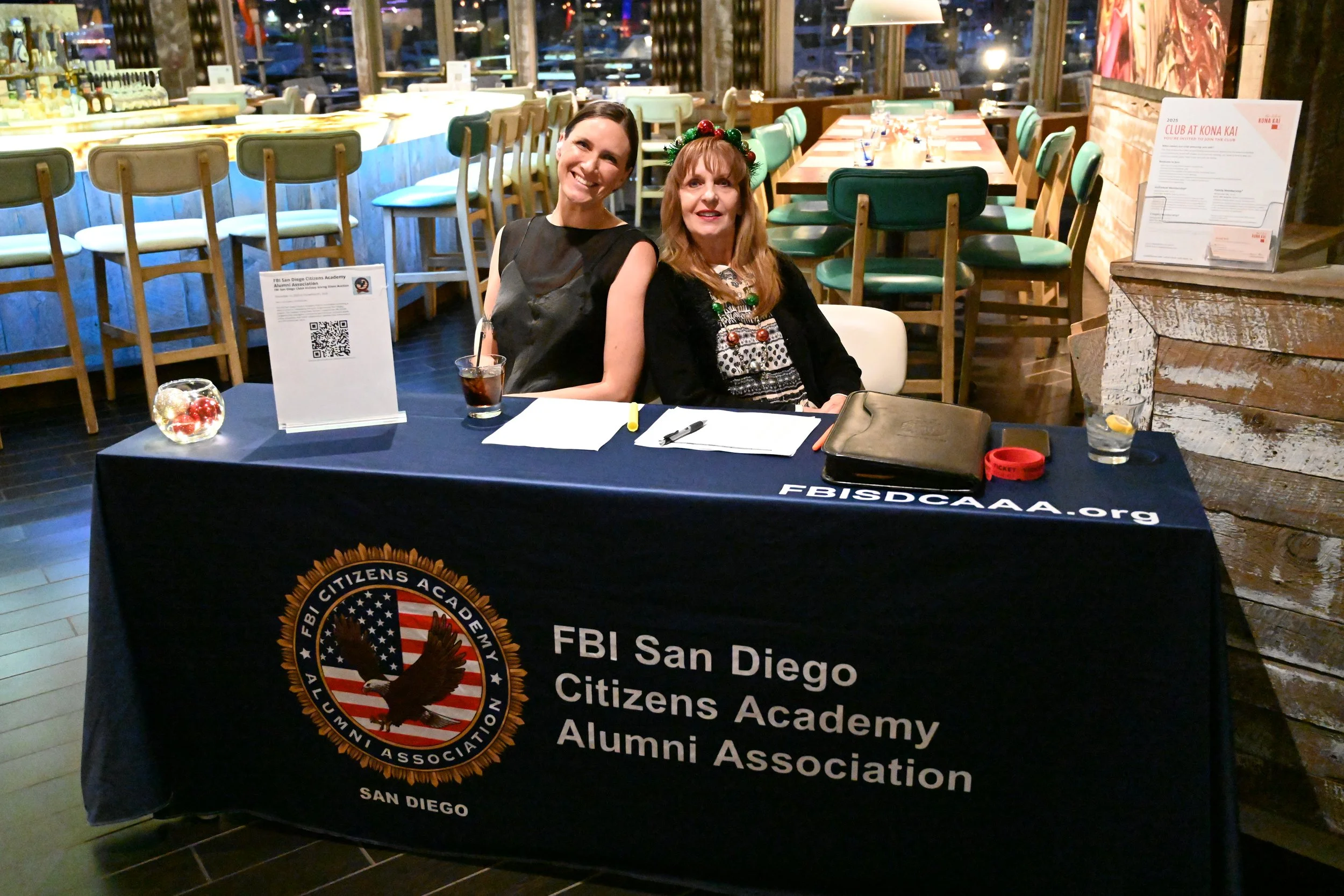 Two women sitting at a table with a black tablecloth that has the text 'FBI San Diego Citizens Academy Alumni Association' and the website 'FBISDCAAA.org'. The woman on the left is smiling, wearing a black sleeveless top, and has short dark hair. The