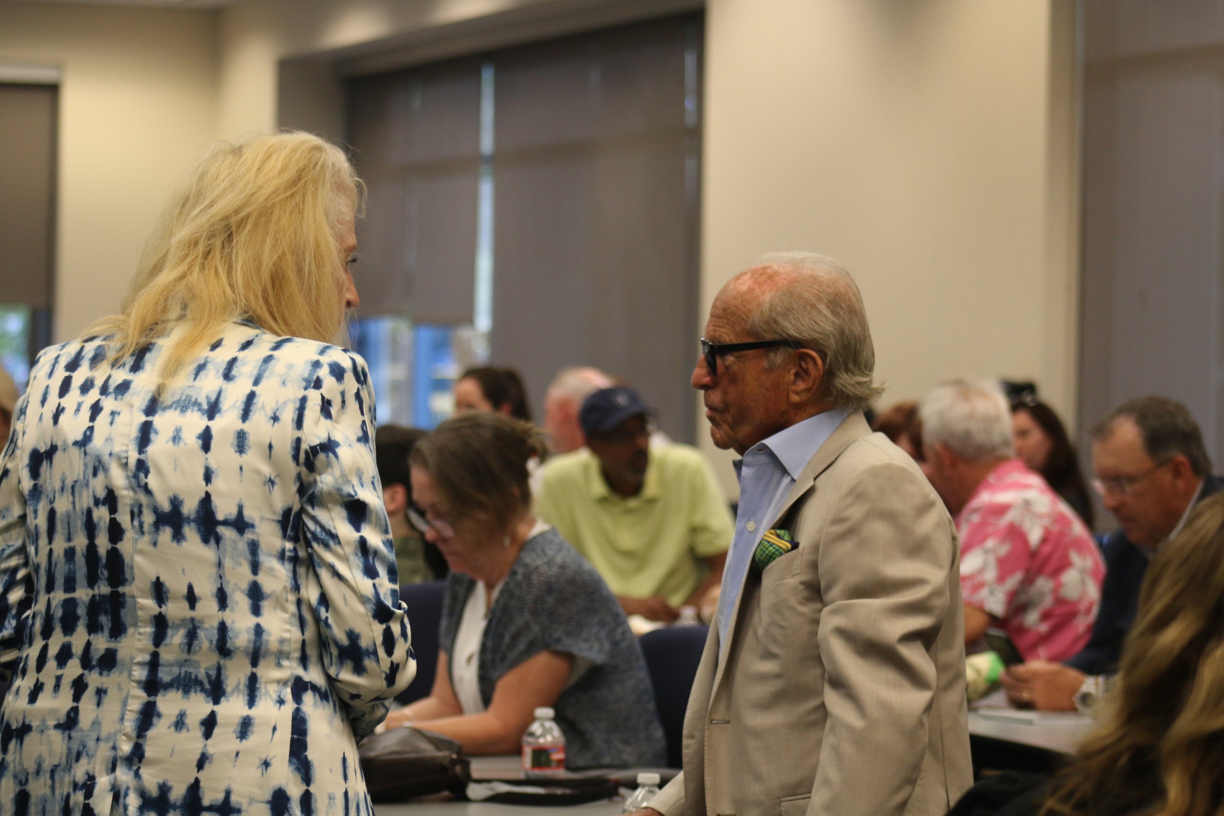 Two people engaged in conversation in a conference room with other attendees seated at tables in the background.
