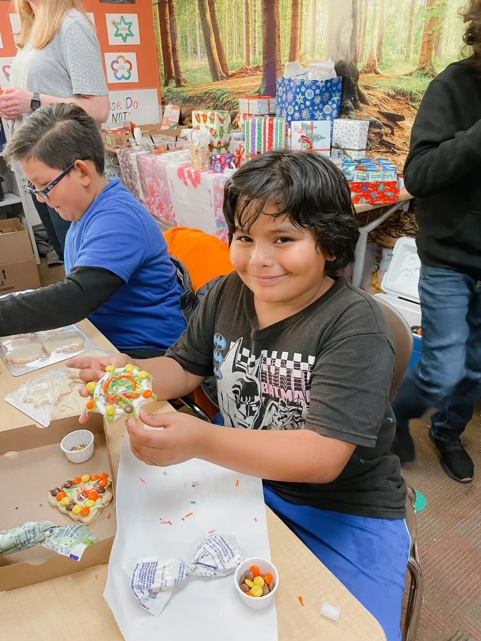 Boy smiling while decorating a holiday cookie with colorful candies in a room decorated for Christmas, with wrapped presents and a woodland-themed backdrop in the background.