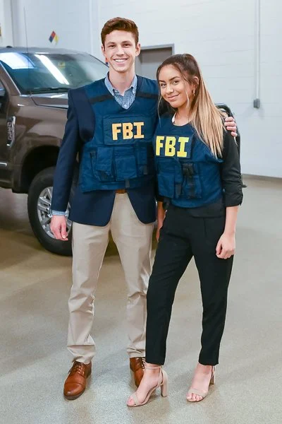 Two FBI agents, a man and a woman, standing and smiling indoors with a vehicle in the background. They are wearing navy blue bureau vests with yellow FBI lettering.