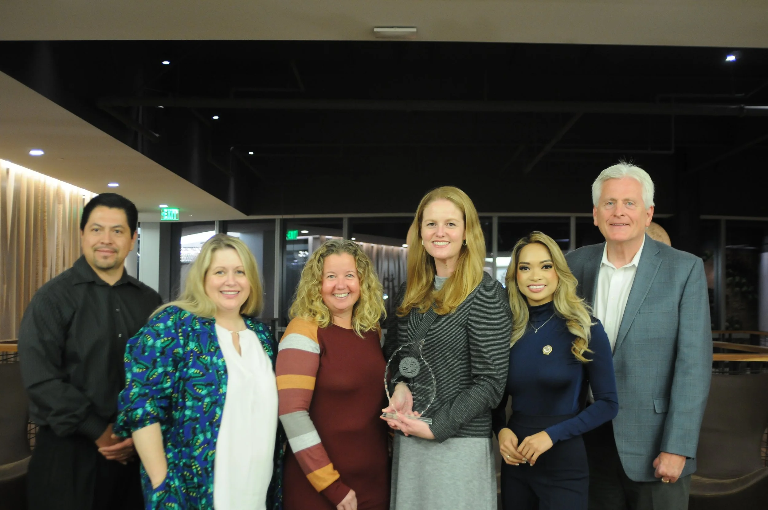 Group of six people smiling, with one woman holding an award, in a modern indoor setting.