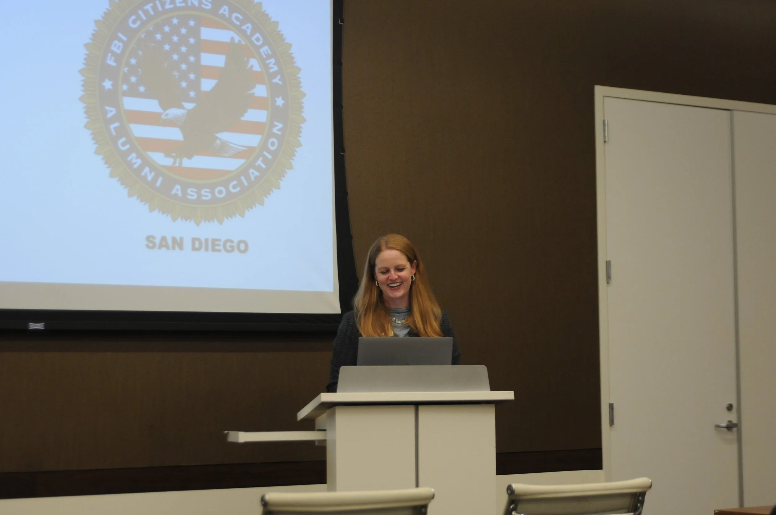 A woman with red hair smiling and looking down at her laptop at a podium, in front of a screen displaying the seal of the FBI Citizens Academy Alumni Association with the words 'SAN DIEGO' underneath.