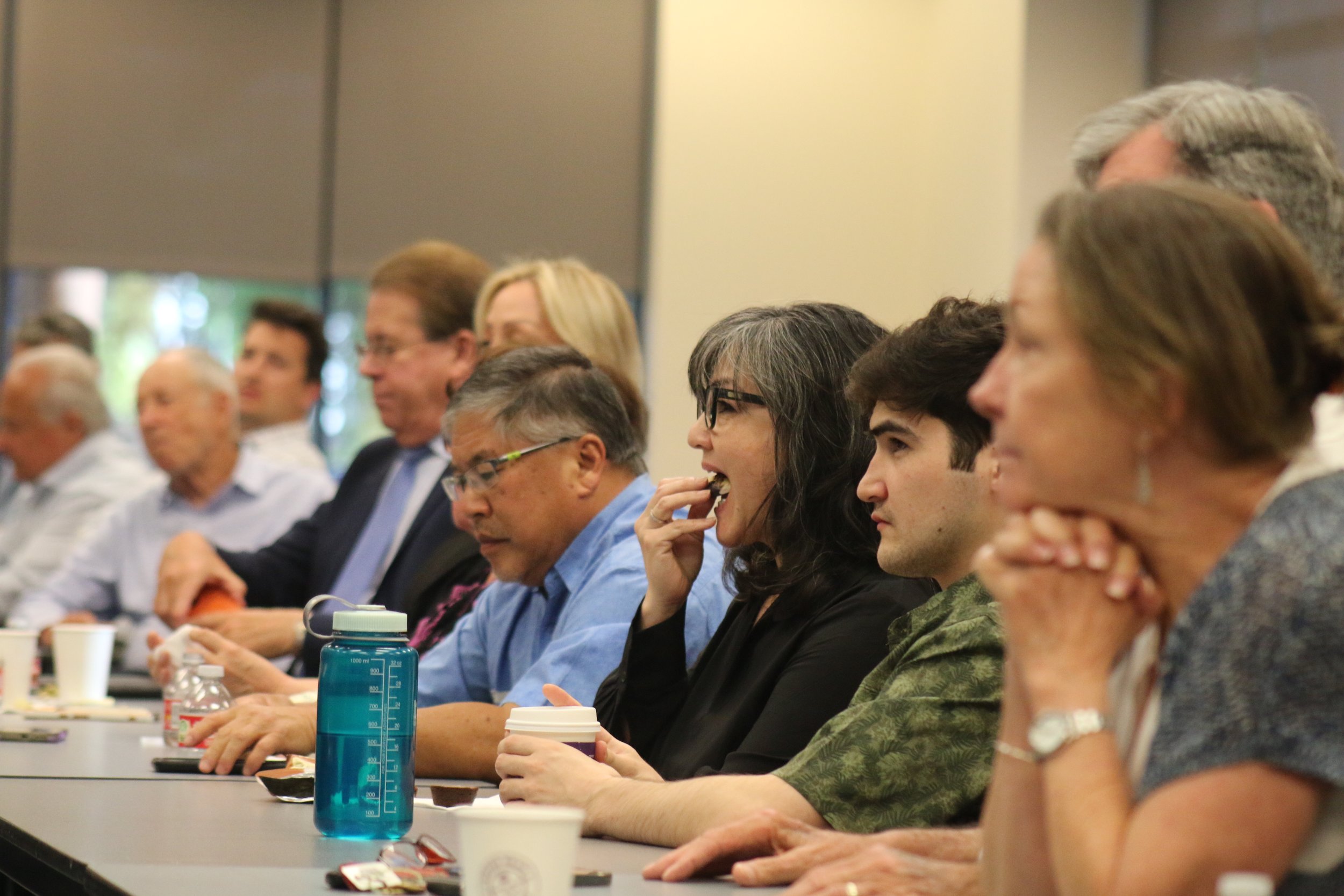 A diverse group of people sitting at a conference table, some drinking coffee, listening attentively, and taking notes during a meeting or seminar.