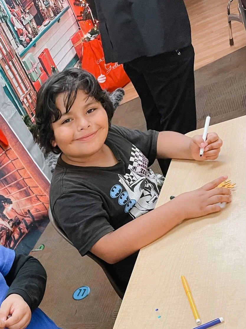 A young boy with curly black hair and a gray T-shirt featuring Star Wars graphics, sitting at a table and smiling at the camera while holding an orange object and a white marker. There are other markers and decorations on the table, and a Christmas d