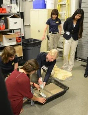 Four women in a room with storage shelves and a trash bin, examining and handling objects in a black tray on the floor, with one woman wearing a blazer and three others in casual clothing.