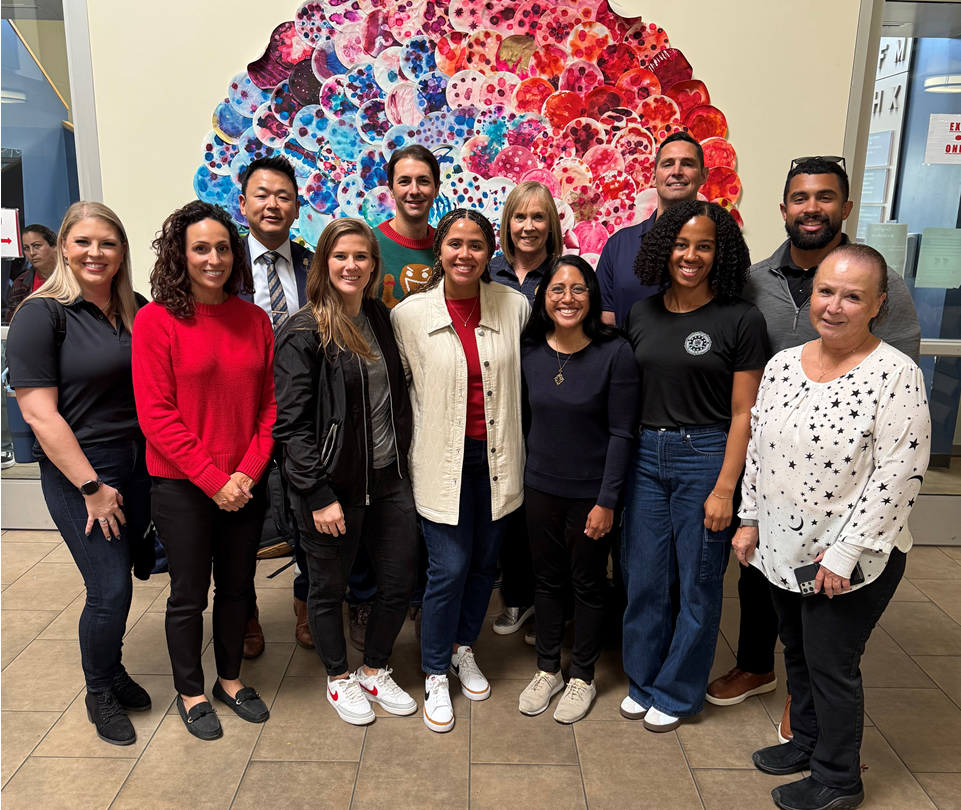 A diverse group of people posing together indoors in front of colorful abstract artwork depicting a large circle of pink, red, purple, and blue dots.