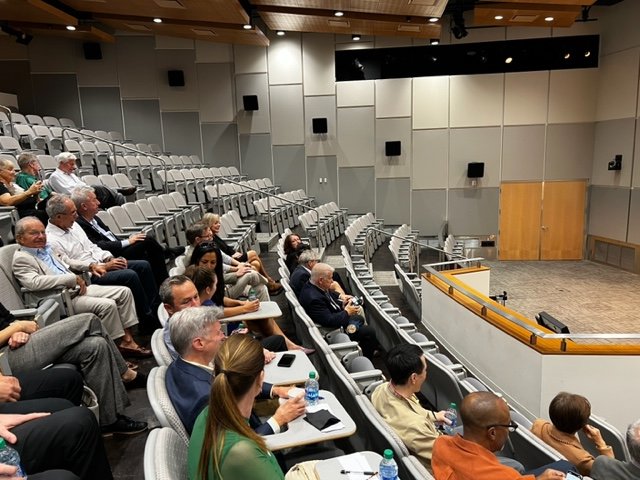 Audience seated in auditorium facing stage with empty podium and screen.
