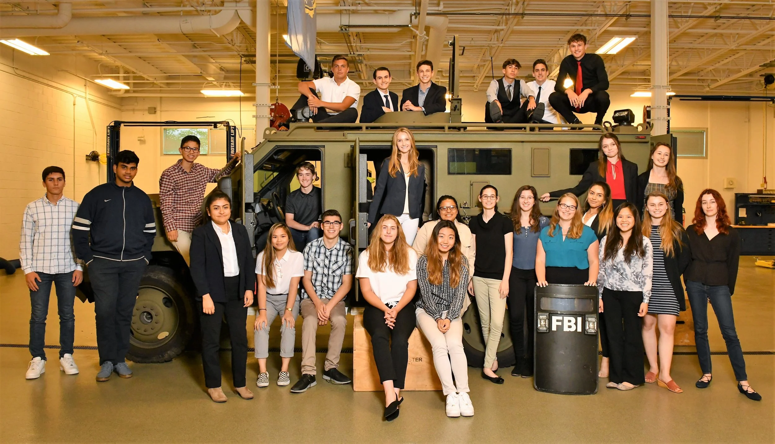 Group of young students and teachers gathered around and on a military-style vehicle inside a school or training facility, with some standing, some sitting on top, and others sitting or standing in front of it, posing for a group photo.