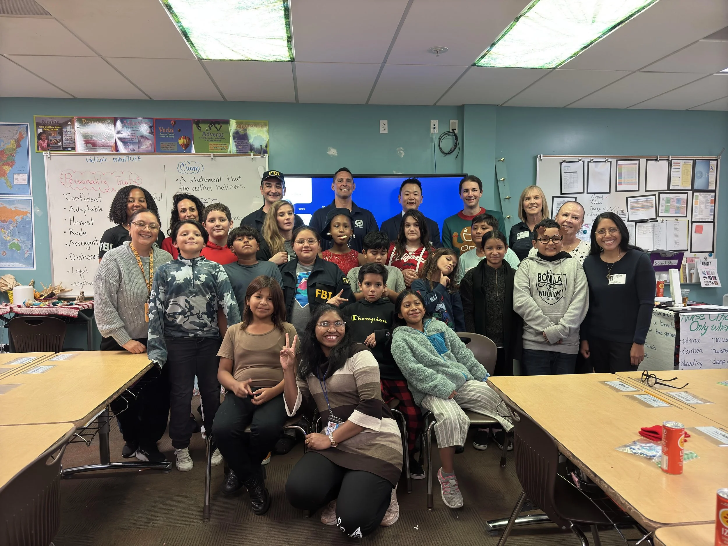 Group of children and adults gathered in a classroom, smiling for a group photo, with whiteboards and educational posters in the background.