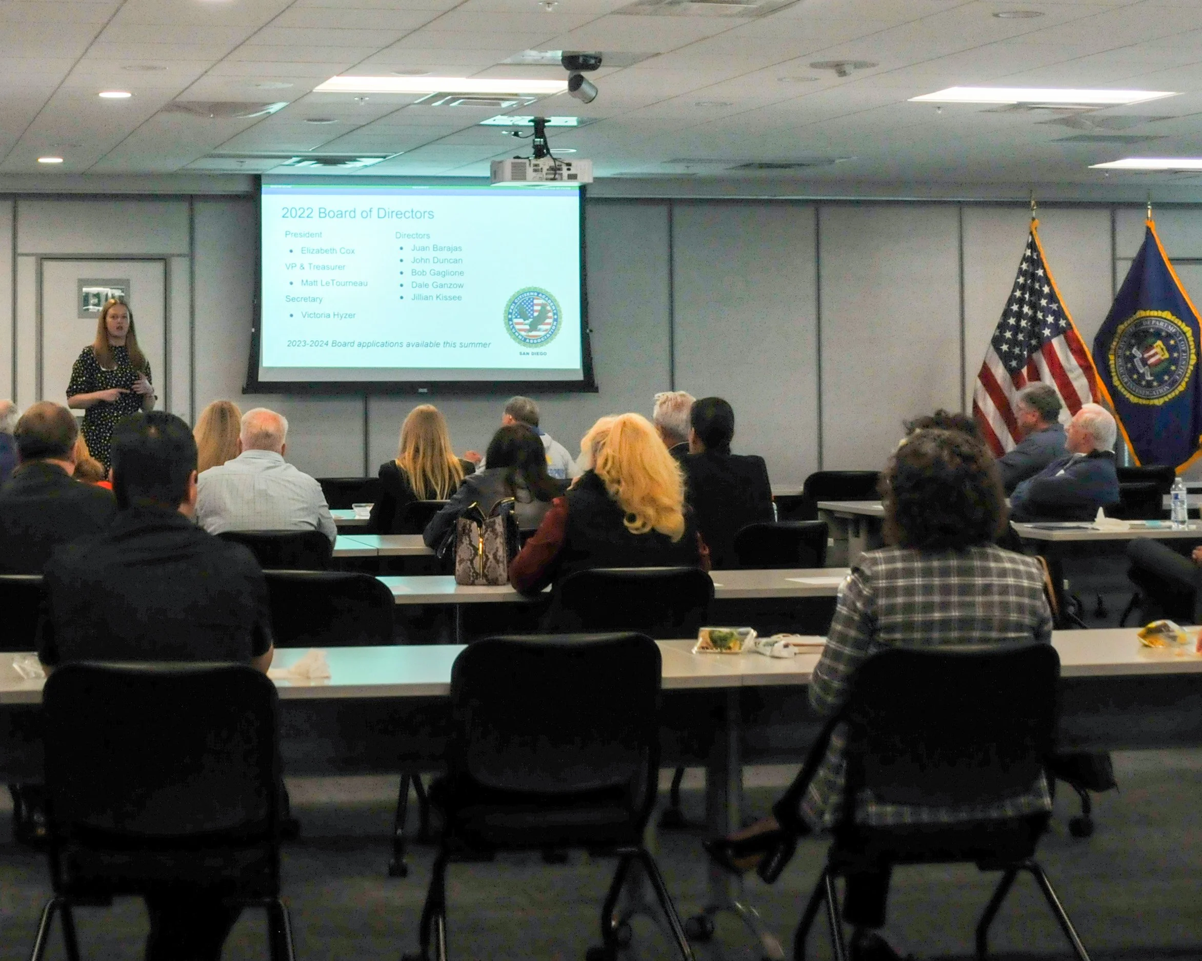 A woman is giving a presentation at a conference room with an audience seated at tables. The slide on the screen displays the 2022 Board of Directors for an organization, with names and titles listed, and flags of the United States and an organization emblem on the right side.