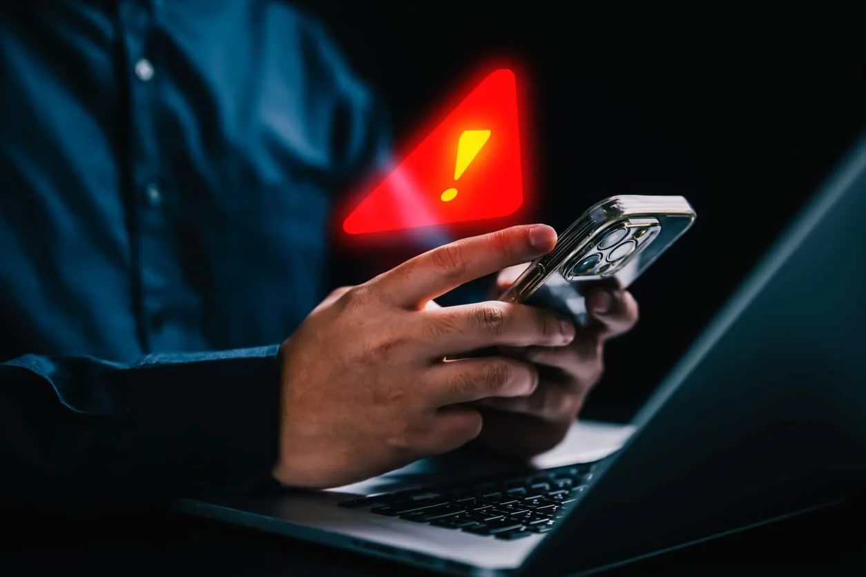 Person holding a smartphone with a warning triangle and exclamation mark above it, in front of a laptop.