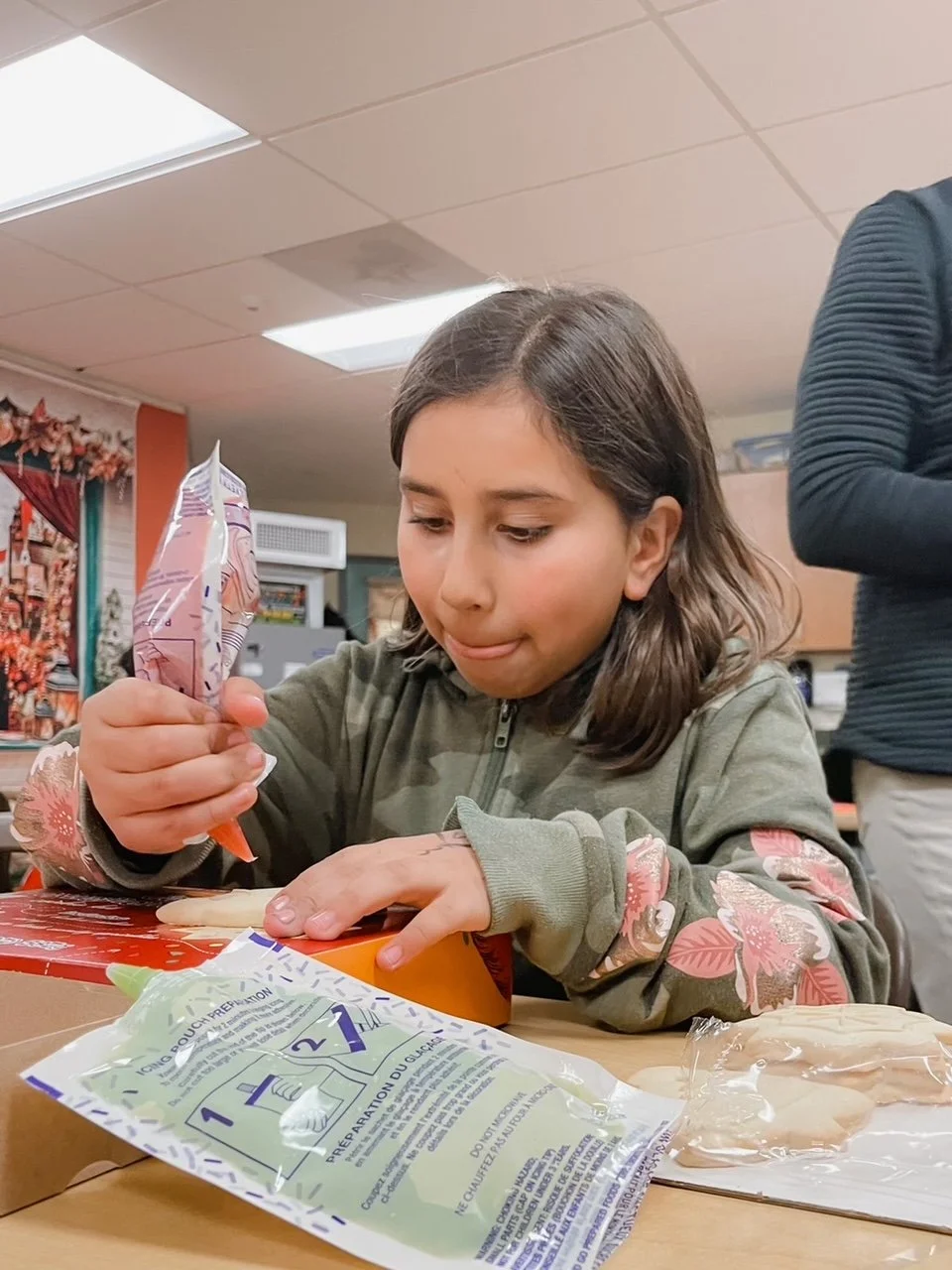 A girl in a gray jacket with floral sleeves is opening a snack package on a table, with a bag of potato chips and a plastic wrapper nearby in an indoor setting.