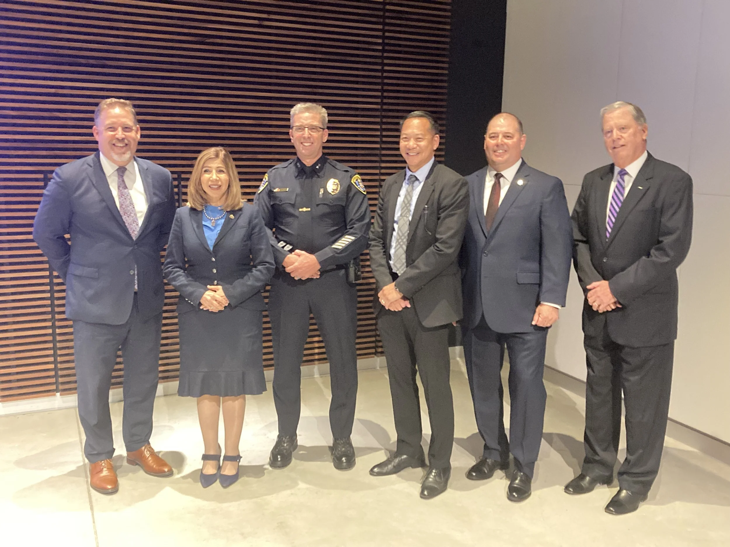 Group of seven professionally dressed individuals, including a police officer, standing together indoors in front of a wooden slat wall.