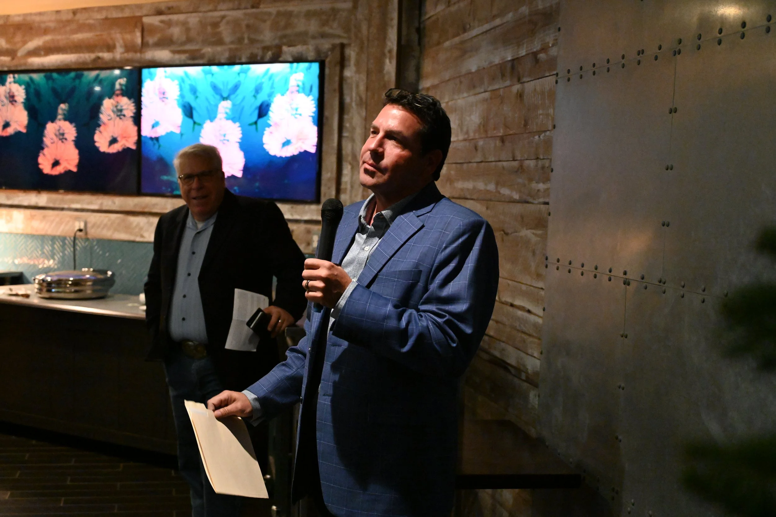 A man in a blue plaid suit jacket holding a microphone and a paper standing in front of a wood-paneled wall, with another man behind him in a dark jacket and light blue shirt, in a dimly lit indoor setting.