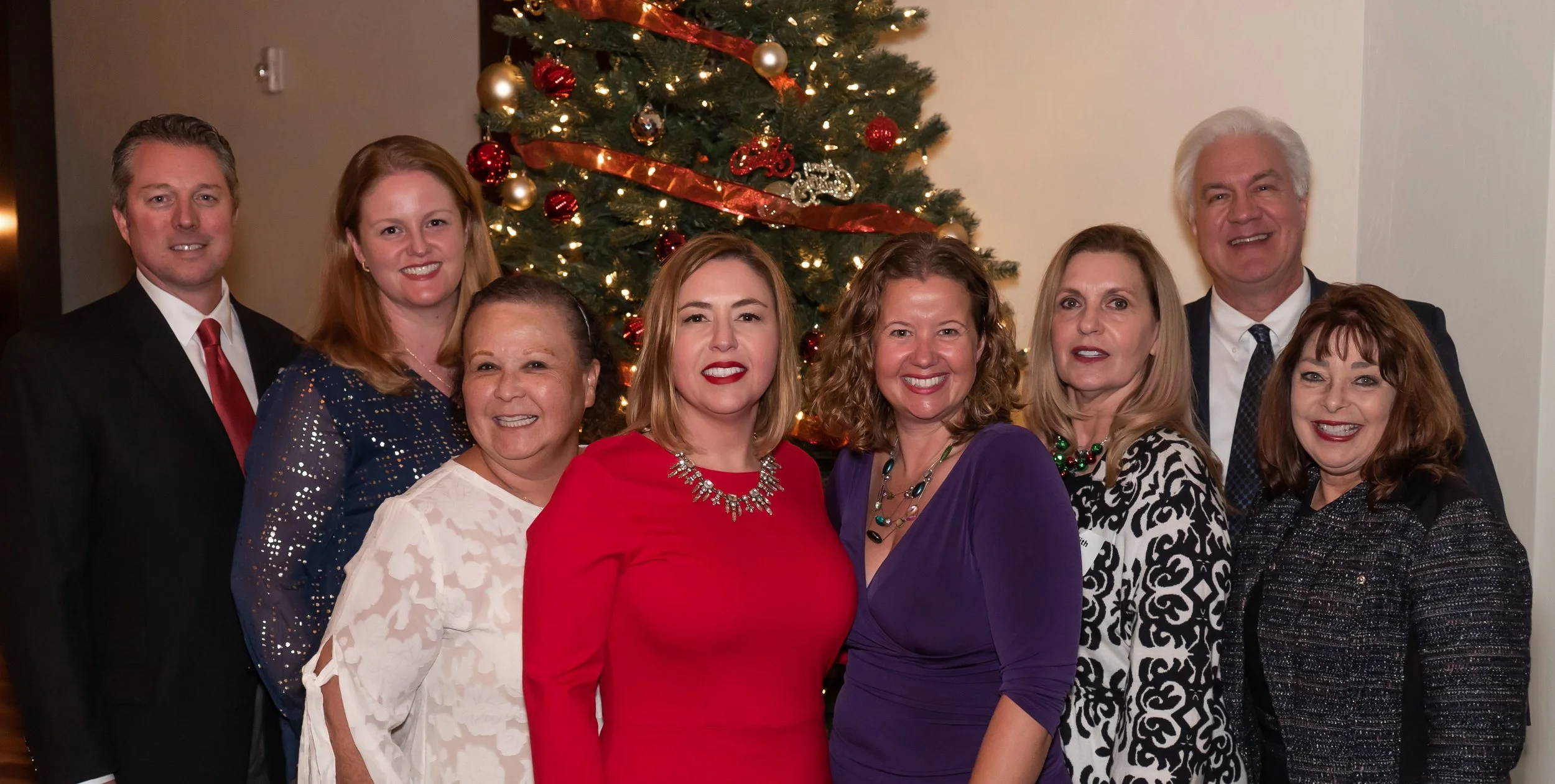 Group of nine people smiling and posing in front of a decorated Christmas tree at a holiday gathering.